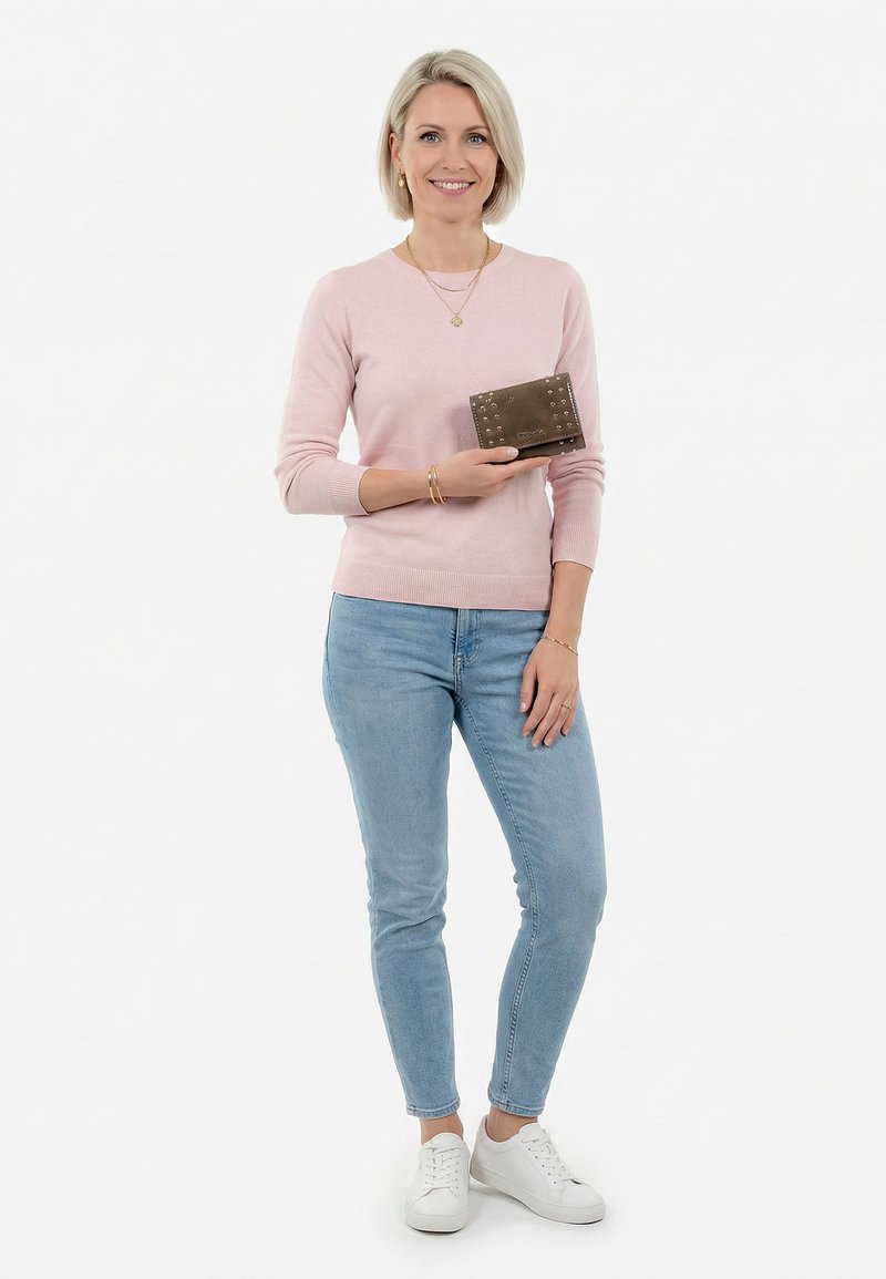 Femme souriante en pull rose et jean tenant un portefeuille marron clouté, debout devant un fond blanc.