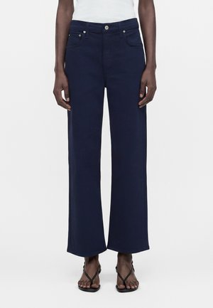 Persona con pantalones anchos de color azul marino, camiseta blanca y sandalias negras con tiras, de pie frente a un fondo claro y liso.