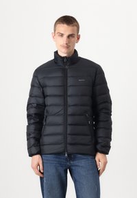 Giacca a vento nera con colletto alto, zip frontale e due tasche laterali con zip. Presenta un design trapuntato e il logo GANT.