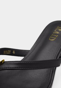 Μαύρο δερμάτινο σανδάλι με λεπτό λουράκι και χρυσή διακοσμητική λεπτομέρεια, κοντινή λήψη στη σόλα που δείχνει το όνομα του εμπορικού σήματος και την ετικέτα με το μέγεθος.