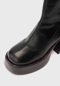 Botín de cuero negro con punta redonda, costuras visibles y un tacón de plataforma grueso y texturizado. Diseño limpio con un ligero brillo.