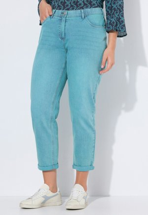 Person trägt hellblaue hochgekrempelte Jeans, weiße Turnschuhe und ein dunkelblaues langärmeliges Oberteil mit Muster, steht mit einer Hand in der Tasche.