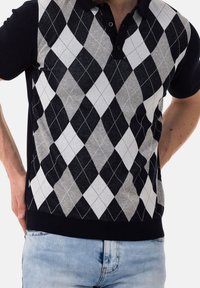 Homme portant un pull à manches courtes noir, gris et blanc à motifs losanges, sur un jean bleu clair, mains posées sur les hanches.