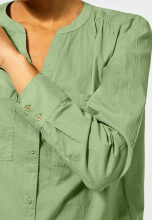 Osoba ubrana w jasnozieloną, długą koszulę bez kołnierzyka, trzymająca lewą rękę prawą dłonią.