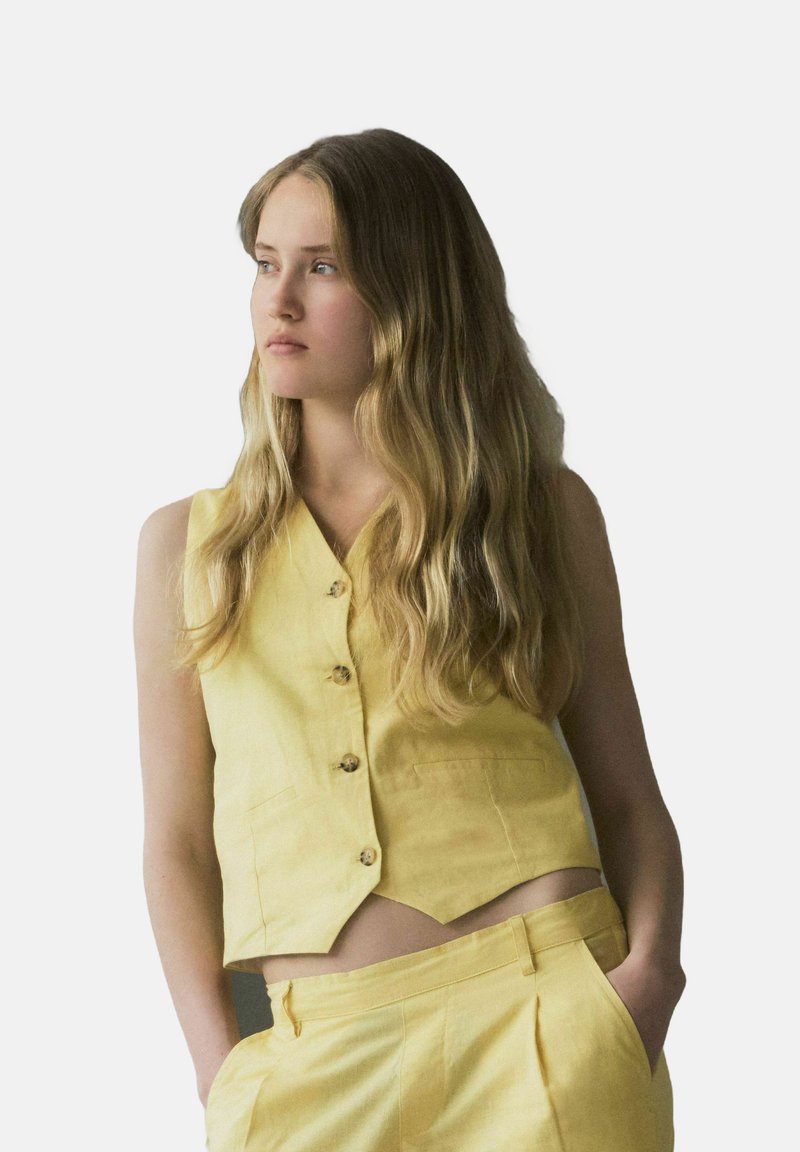 Chaleco de lino amarillo con escote en forma de V, cinco botones y dos bolsillos; combinado con pantalones a juego, con cintura ajustada y pinzas.