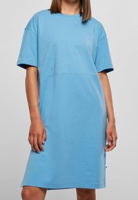 Niebieska bawełniana sukienka koszula z krótkimi rękawami, luźny krój, długość do kolan i mała zielona metka z boku. Gładka tekstura, minimalistyczny design.