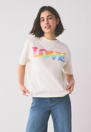 Junge Frau mit kurzen dunklen Haaren, die ein weißes T-Shirt mit dem regenbogenfarbenen Schriftzug "LOVE" und blaue Jeans trägt, vor einem schlichten Hintergrund stehend.