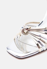 Ασημένιο πέδιλο με ανοιχτά δάχτυλα και ψηλό τακούνι, με μεταλλική λουρίδα με κόμπο και τετράγωνη μπροστινή σόλα.