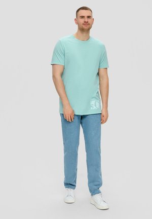 Lichtblauwe katoenen t-shirt met korte mouwen, ronde halslijn en een subtiele grafische detail aan de zijkant, gecombineerd met lichtblauwe jeans en witte sneakers.