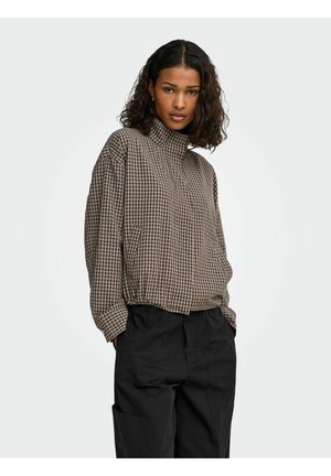Femme aux cheveux bouclés portant un haut à manches longues marron à carreaux et un pantalon noir ample, les mains dans les poches, regardant vers l'avant.