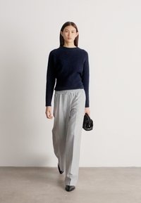 Sieviete pastaigā telpās, valkājot melnu džemperi, vaļīgas gaiši pelēkas bikses, melnus smailus apavus, tur mazs melns rokassomas.