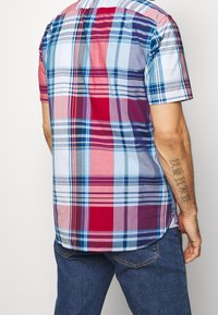 Camicia a maniche corte a quadri con toni rossi, blu e bianchi, disegni a controllo vario, orlo arrotondato e dettagli di cucitura sottili sul retro.