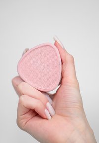 Różowa silikonowa szczotka w kształcie trójkąta z textured powierzchnią z drobnymi okrągłymi wzorami, trzymana w dłoni na szarym tle.