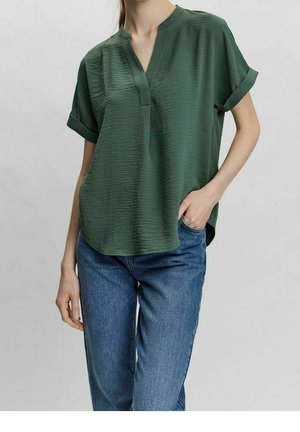 Mujer con blusa verde de manga corta y holgada, con los puños doblados, y vaqueros azules, con una mano en el bolsillo, de pie contra un fondo liso.