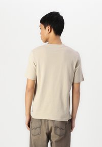 Camiseta de algodón beige de manga corta, con cuello redondo y un ajuste relajado, combinada con pantalones de mezclilla marrón claro que presentan costuras en los bolsillos traseros.