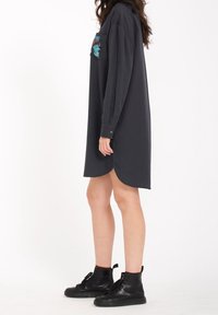 Robe chemise gris charbon à manches longues, fentes sur les côtés et un patch poitrine avec un motif brodé coloré. Portée avec des bottines.