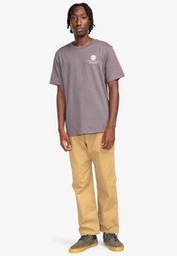 Grijze katoenen T-shirt met een logo op de borst, gecombineerd met tan broek en groene sneakers. De algehele pasvorm is ontspannen en casual.