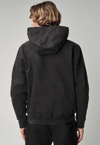 Hanorac negru cu o glugă mare, cu șnur, manșete și tiv cu nervuri. Material neted cu detalii de cusătură, purtat împreună cu pantaloni dark asortați.