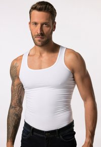 Hvid tanktop lavet af bomuld, der strækker sig over en muskuløs bygning, komplementeret af mørkeblå denimjeans og synlige tatoveringsdetaljer på armen.