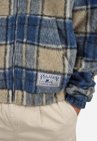 Fleece zadrgana jakna v modro-bež karirastem vzorcu z teksturirano površino. Opremljena je z logotipom, ki pravi "PEGADOR Everyday Ambition."
