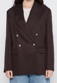 Femme portant un blazer marron foncé à double boutonnage avec quatre boutons et des poches à rabat à l'avant, par-dessus une chemise claire et un jean bleu.