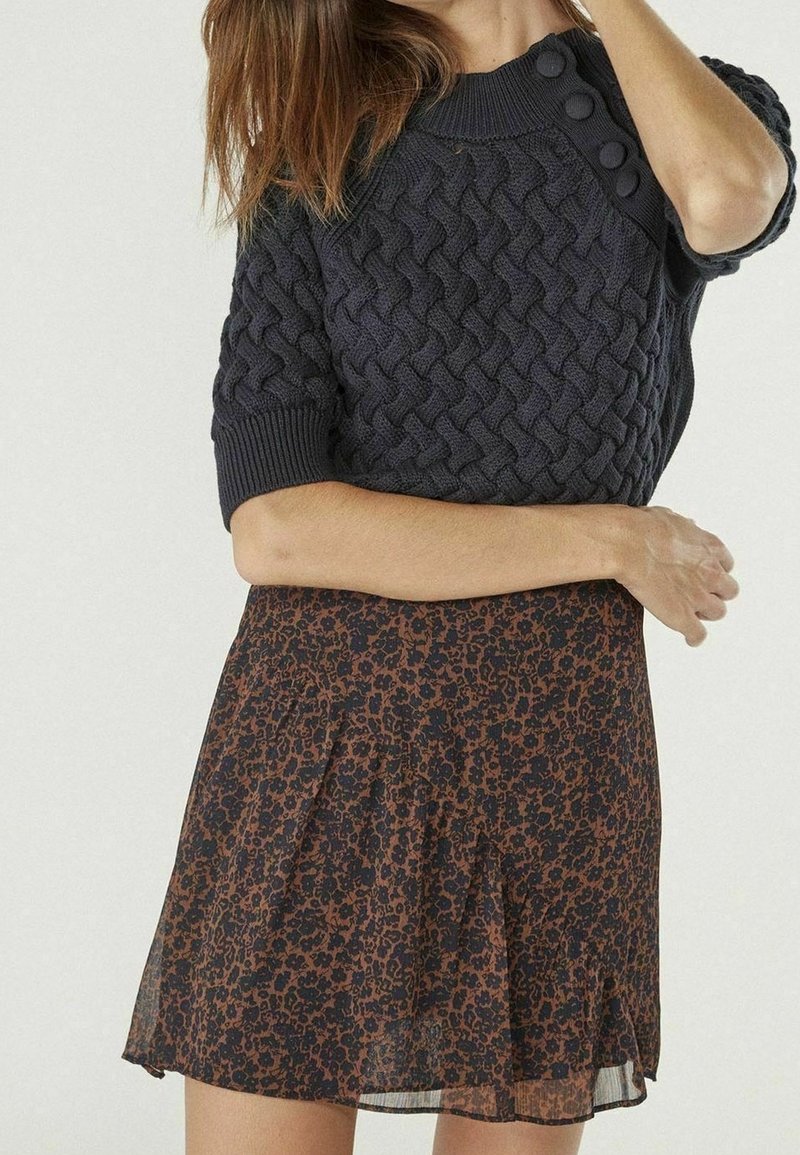 Vrouw die een donker kabelgebreide korte mouwtrui en een bruine rok tot op de knie met een zwart bloemenpatroon draagt.