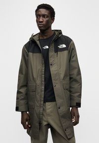 Αδιάβροχο μπουφάν παρκά σε ελιά, με μαύρο επάνω τμήμα και κουκούλα, διαθέτει διπλά λογότυπα, εμπρόσθιες τσέπες και κλείσιμο με κουμπιά πατιλέτα.