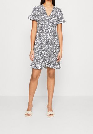 Vrouw draagt een korte zwarte wikkeljurk met witte bloemenprint, vlindermouwen, een gerimpelde zoom en sandalen met gevlochten bandjes, staand tegen een witte muur.