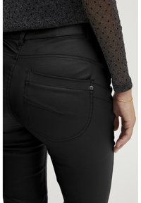 Personne portant un pantalon noir ajusté avec des détails de poches arrière et un haut transparent à manches longues avec un motif à pois, la main reposant sur le côté.