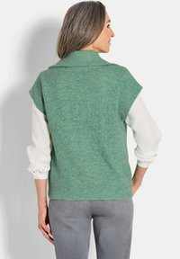 Pull sans manches en tricot côtelé vert avec un col haut, porté sur une chemise blanche à manches longues, associé à un jean gris.