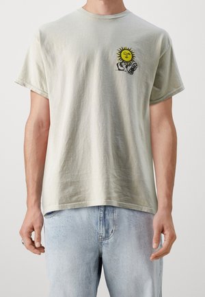Lysegrøn bomuld t-shirt med en grafik af en gul sol og hænder, korte ærmer og en afslappet pasform. Båret med lyseblå jeans.