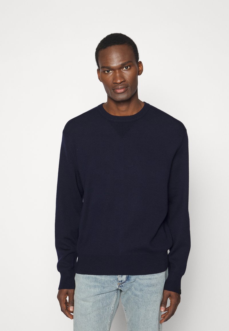 Pull en maille bleu marine avec un col rond, des poignets et un ourlet côtelés, présentant un motif texturé au niveau des épaules. Modèle portant un jean bleu clair.
