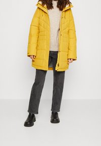 Person som bär en gul puffjacka över en beige tröja, mörkgrå jeans och svarta ankelboots, som står mot en neutral bakgrund.