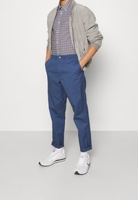 Ljusgrå bomberjacka över en rutig skjorta i blått och vitt, ihop med marinblå byxor och vita sneakers med svarta accenter.