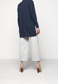 Camicia di denim blu scuro, maniche lunghe, design con piega sul retro. Abbinate a pantaloni larghi in beige chiaro e scarpe con tacco bianche e cinturini.
