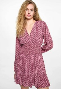 Short dress in patterned pink fabric with a smocked waist, long sleeves, V-neckline, and ruffled hem. Features small floral designs.