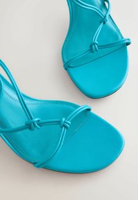 Sandales turquoise à bout ouvert avec fines brides nouées et semelles carrées sur un fond neutre.