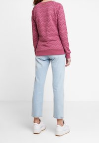 Persona vestida con una camiseta de manga larga de patrón de chevron rosa, jeans cortos de color azul claro y zapatillas blancas, de pie contra un fondo blanco.