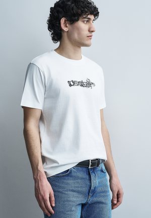 Νεαρός άνδρας με σγουρά μαλλιά που φοράει λευκό t-shirt Off-White και μπλε τζιν, στέκεται μπροστά από απλό γκρι ανοιχτό φόντο.