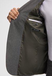 Blazer grigio con un esterno testurizzato e un interno liscio verde oliva; presenta una tasca laterale e un etichetta che dichiara "MIGLIOR TESSUTO ITALIANO".