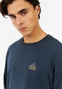 Marineblauwe lange mouwen t-shirt van katoen, met een klein gouden driehoeklogo op de borst. Soepele textuur en ronde halslijn.
