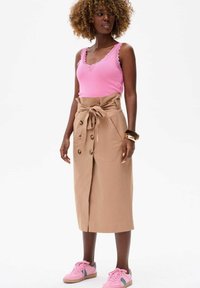 Débardeur en tricot rose clair avec des bords en scaloppé, associé à une jupe tan longueur genoux avec une taille à nouer et des détails de boutons sur le devant, complété par des baskets roses.