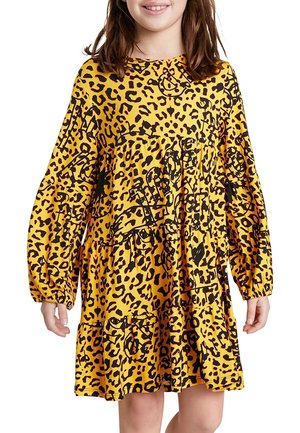 Dziecko ubrane w żółtą sukienkę z długim rękawem, ozdobioną czarnymi abstrakcyjnymi i lampartowymi wzorami, stojące z ramionami swobodnie opuszczonymi wzdłuż ciała.