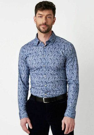 Mann trägt ein eng anliegendes blaues Hemd mit Paisley-Muster, schwarzen Gürtel und dunkle Hose, steht vor einem schlichten weißen Hintergrund.