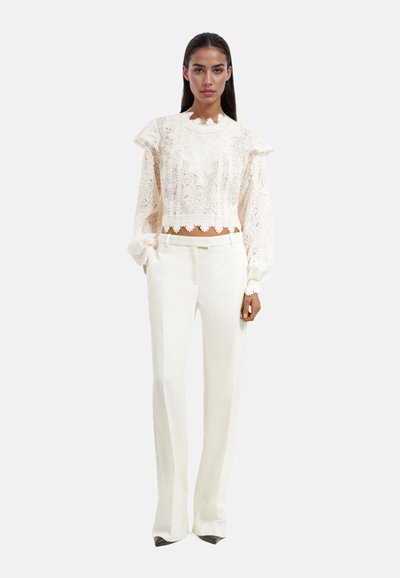 Kanten blouse met ruffles op de mouwen, een gegolfde zoom en hoge hals. Gepaard met op maat gemaakte witte broeken en zwarte puntige hakken.