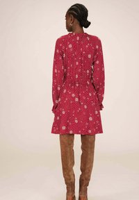 Robe fleurie rouge à manches longues, taille froncée et ourlet évasé. Présente un motif texturé de fleurs blanches et de lignes courbées.