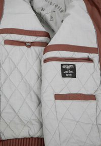 Gepolstertes graues Innenfutter der Jacke mit braunen Lederausschnitten, das über mehrere Taschen und ein Pflegeetikett verfügt. Die Textur ist glatt und gepolstert.