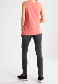 Person i en ärmlös koralltop, mörkgrå skinny jeans och svarta sneakers som står framför en enfärgad vit bakgrund.