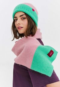 Pletená čepice v zelené a růžové barvě, spárovaná s růžovým a zeleným šátkem. Oba kusy mají jemnou texturu a štítek s názvem značky.