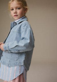 Randig jeansjacka i blå och vita nyanser, med framfickor, bärs över en ljusblå och rosa randig skjorta, vilket fångar en lagerstil.